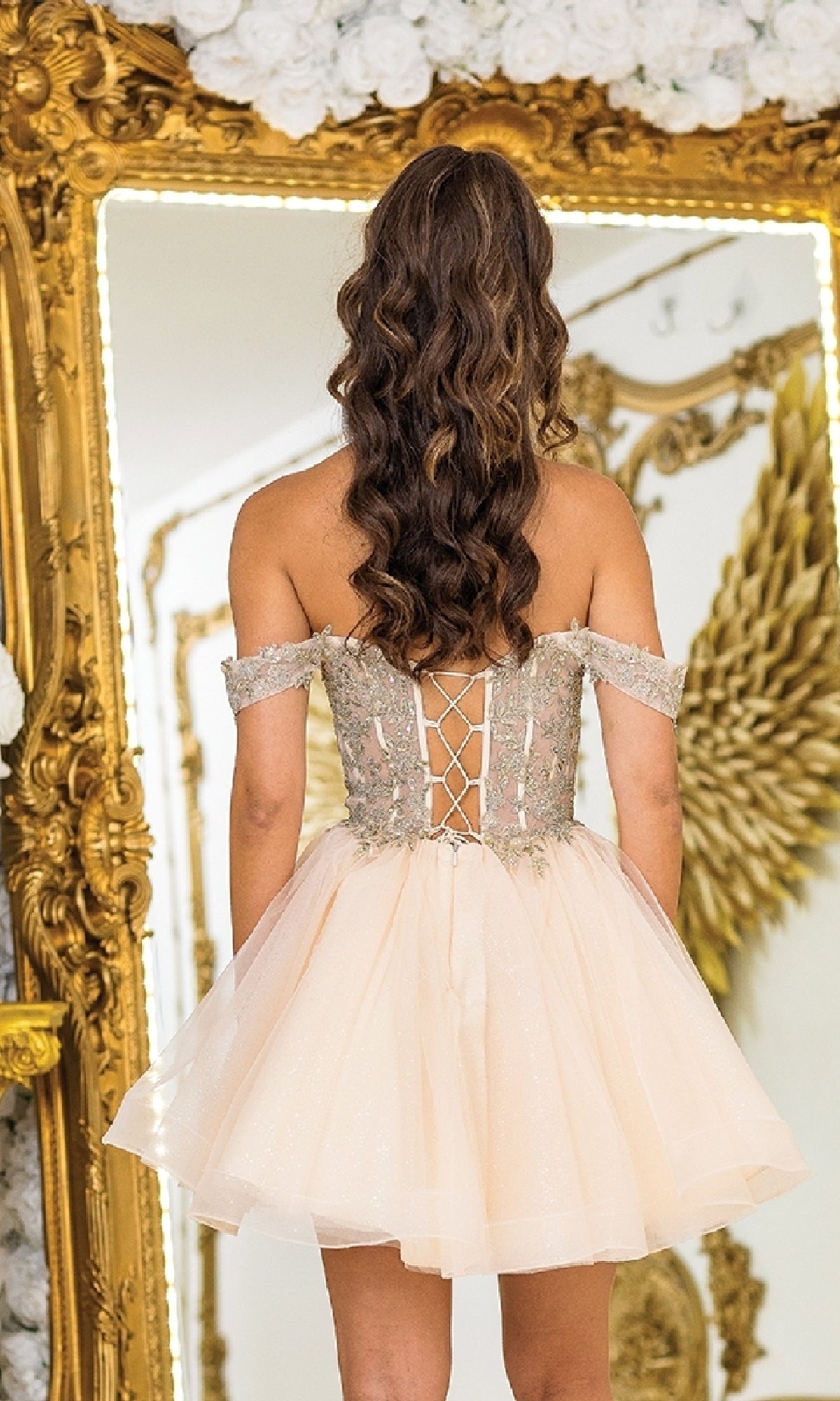 A woman with wavy brown hair stands before an ornate mirror, wearing the Off-Shoulder Short Prom Dress: Dancing Queen 3418—with a lace-up back and tulle skirt—ideal as an affordable prom or babydoll party dress.